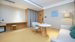 Ein modernes Hotelzimmer mit einem runden Holztisch, einem weißen Sofa, einem kleinen Hocker, einer Minibar und einer Deckenleuchte.