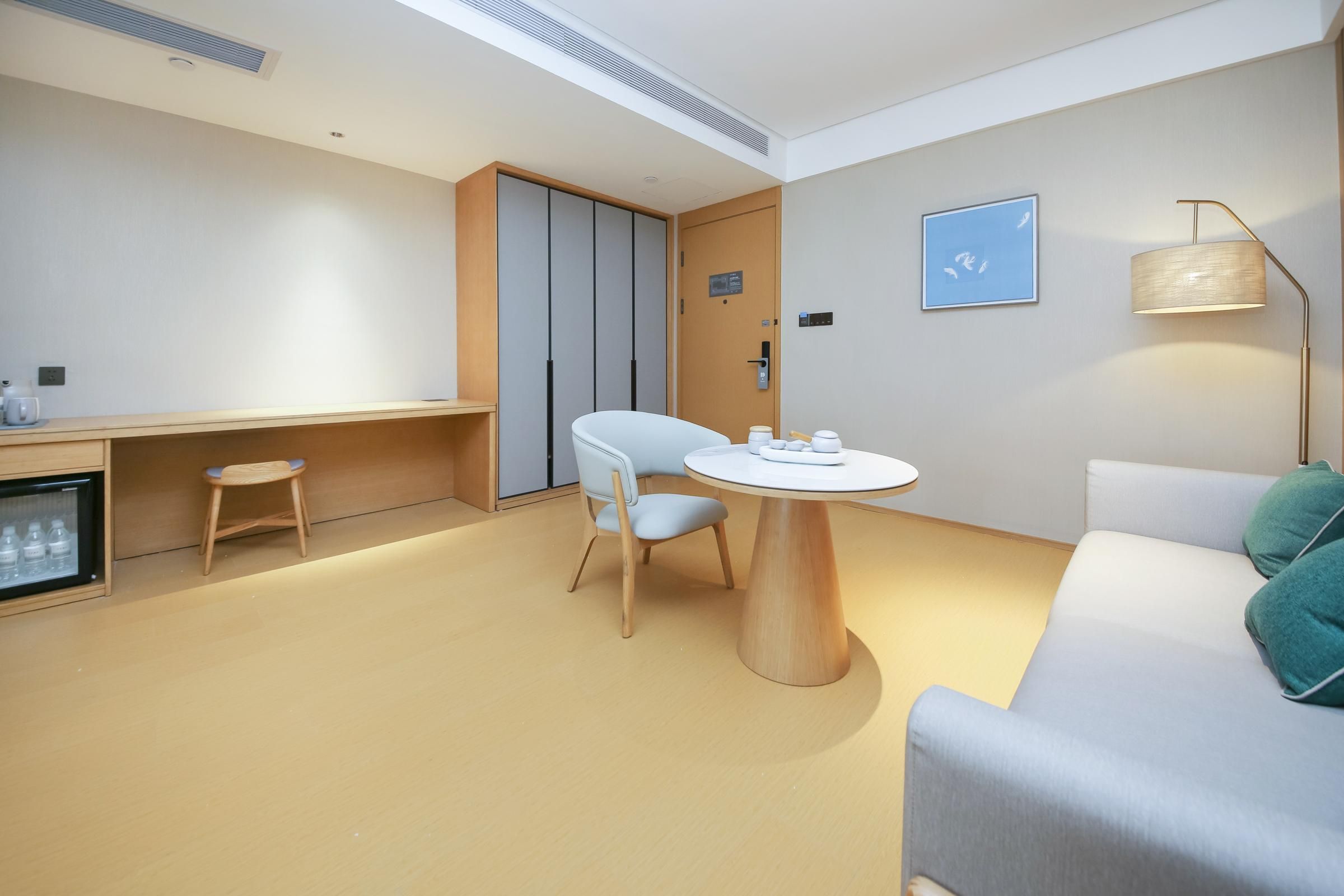 Una habitación de hotel moderna con una mesa redonda de madera, un sofá blanco, un taburete pequeño, un minibar y una lámpara en el techo.