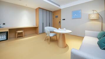 Una habitación de hotel moderna con una mesa redonda de madera, un sofá blanco, un taburete pequeño, un minibar y una lámpara en el techo.