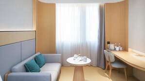Ein modernes Hotelzimmer mit Sofa, einem kleinen runden Tisch, einem Schreibtisch und einem Fenster mit Vorhängen.