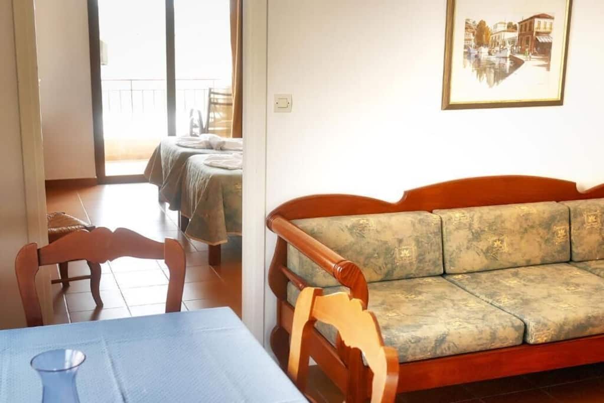 A room with a wooden sofa, a dining table with a vase, and a balcony with a view of buildings.