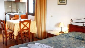 Habitación de hotel con cama, un pequeño escritorio con lámpara, una silla, una mesa con un florero y una cocina visible a través de una puerta abierta.