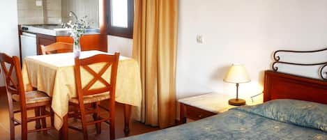 A hotel room with a bed, a small desk with a lamp, a chair, a table with a vase, and a kitchen area visible through an open door.