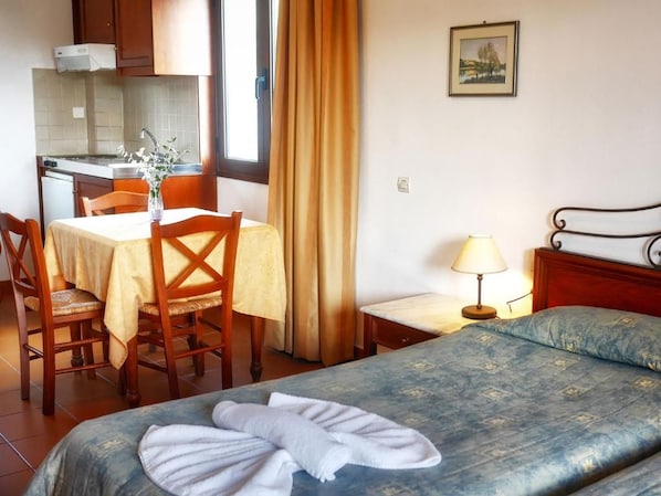 A hotel room with a bed, a small desk with a lamp, a chair, a table with a vase, and a kitchen area visible through an open door. - Areti Apart Hotel (Chania)