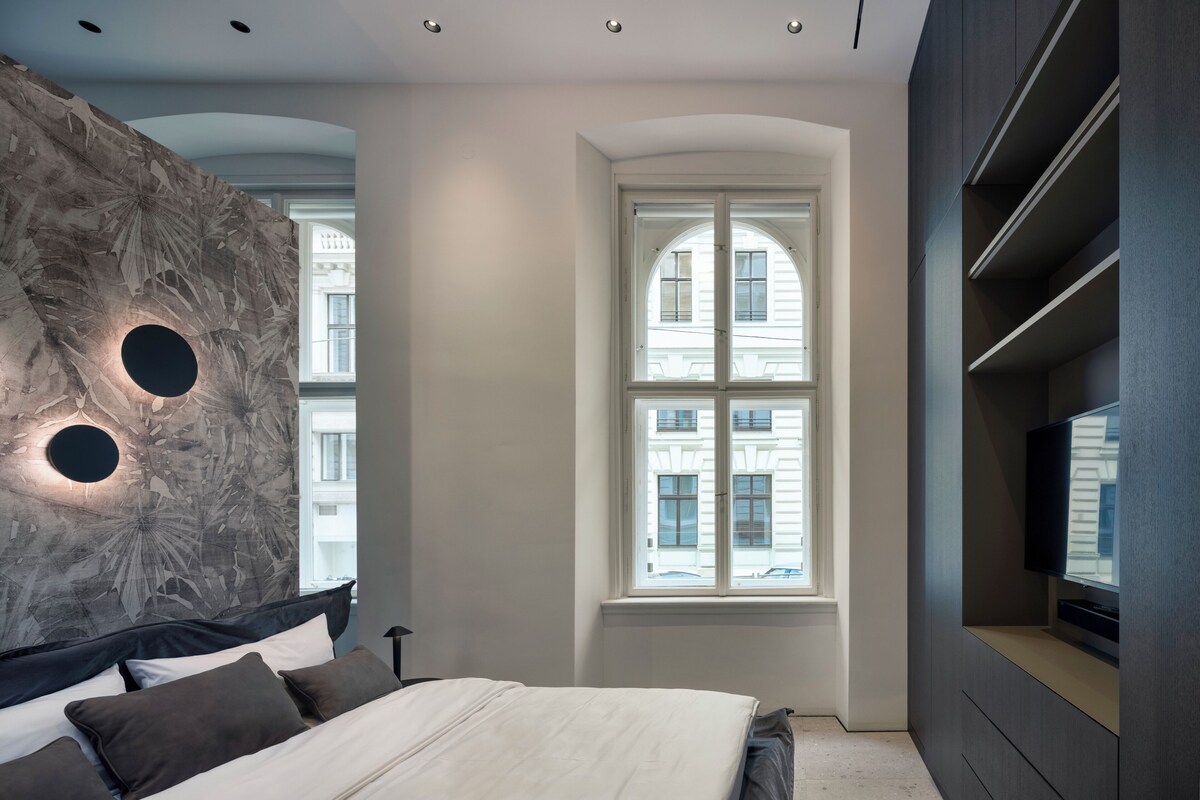 Ein modernes Schlafzimmer mit einem großen Bett, einem an der Wand montierten Fernseher und einem Fenster mit Blick auf Gebäude.