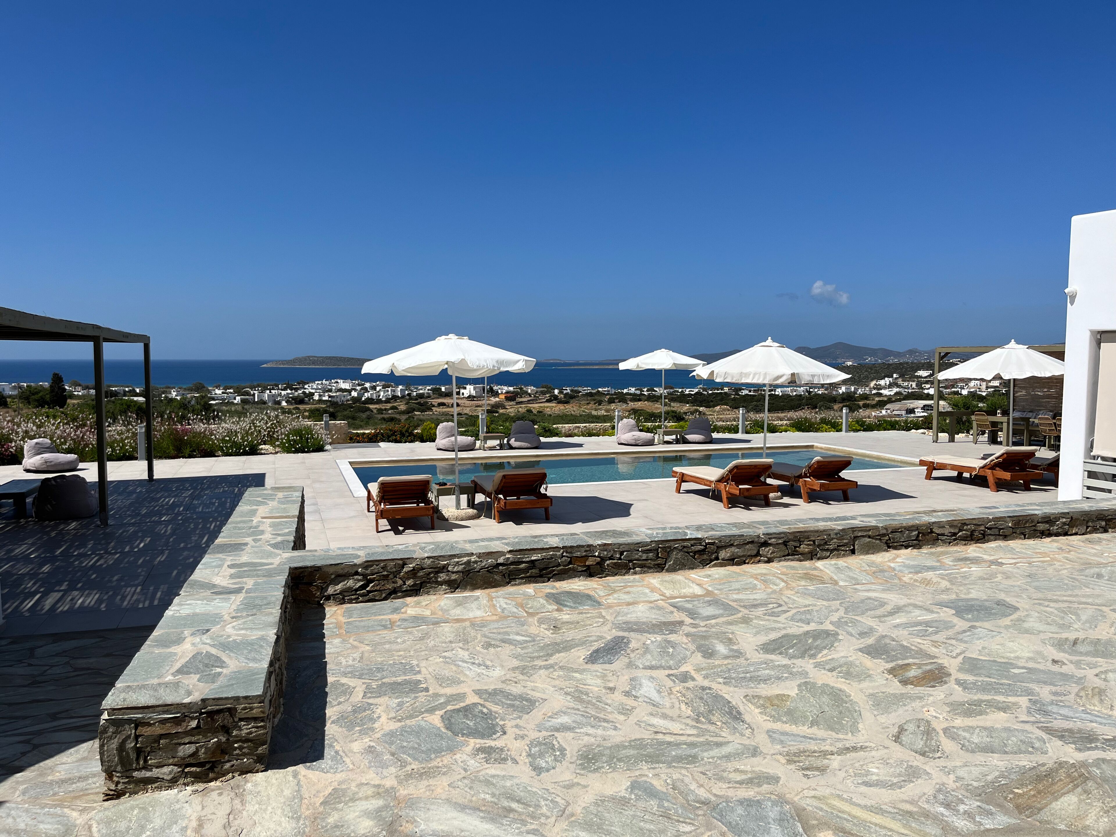 A pool area with lounge chairs and umbrellas, a stone-tiled deck, and a view of the sea and distant buildings.