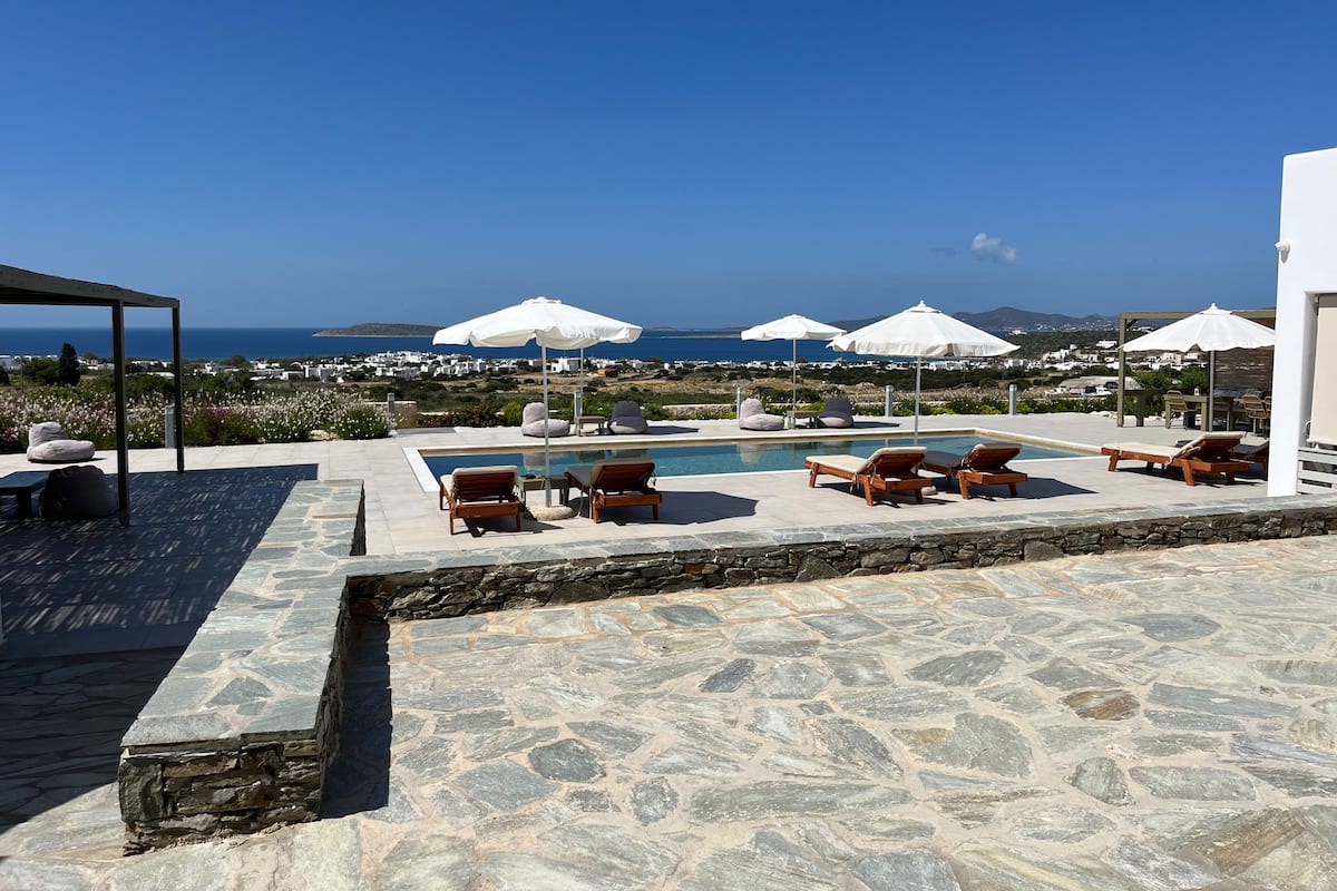 Ein Poolbereich mit Liegestühlen und Sonnenschirmen, eine terrasse mit Steinplatten und Blick auf das Meer sowie entfernte Gebäude.