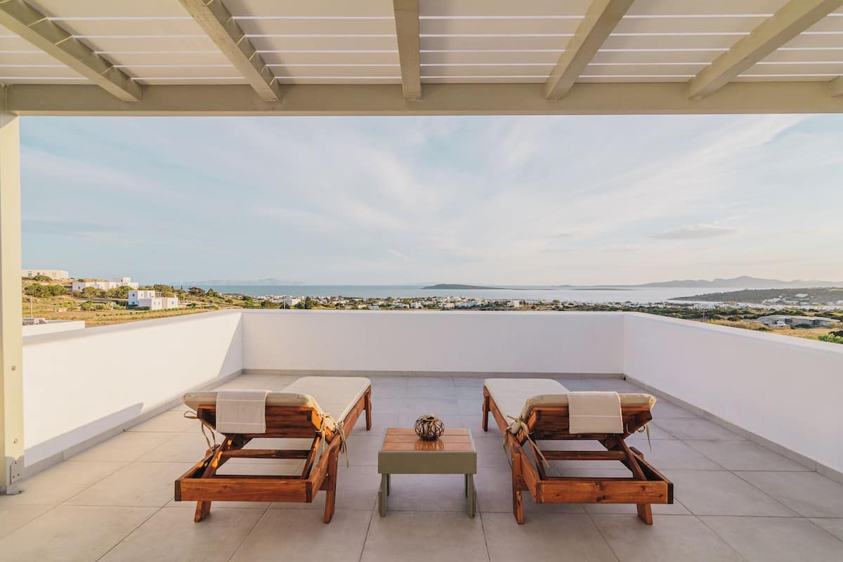 Eine überdachte Terrasse mit zwei Holzliegen, einem kleinen Tisch und Blick auf eine Küstenlandschaft.