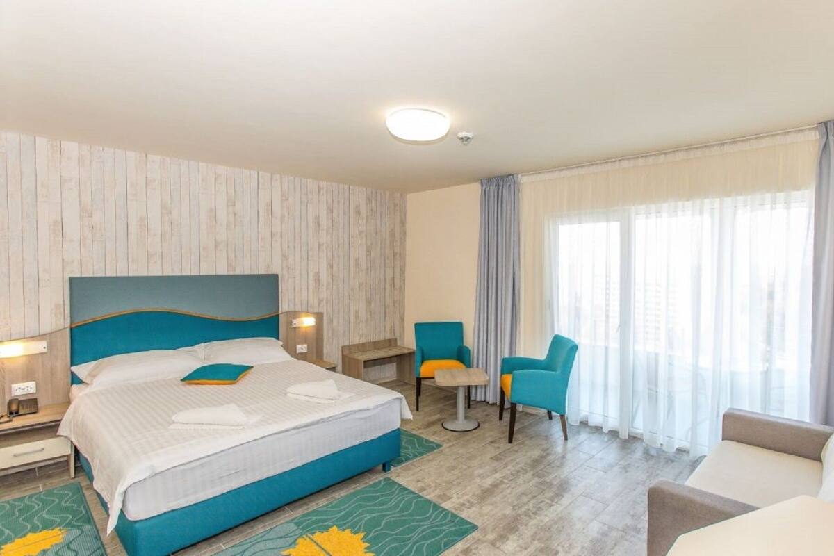 Ett hotellrum med en säng, ett skrivbord, en stol, en soffa och ett stort fönster med gardiner.