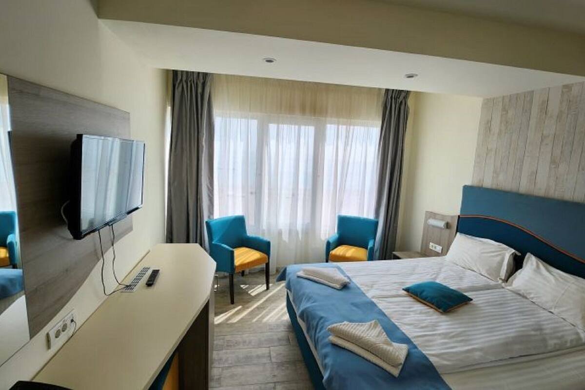 Ett hotellrum med en säng, två stolar, ett skrivbord, en tv och ett fönster med gardiner.