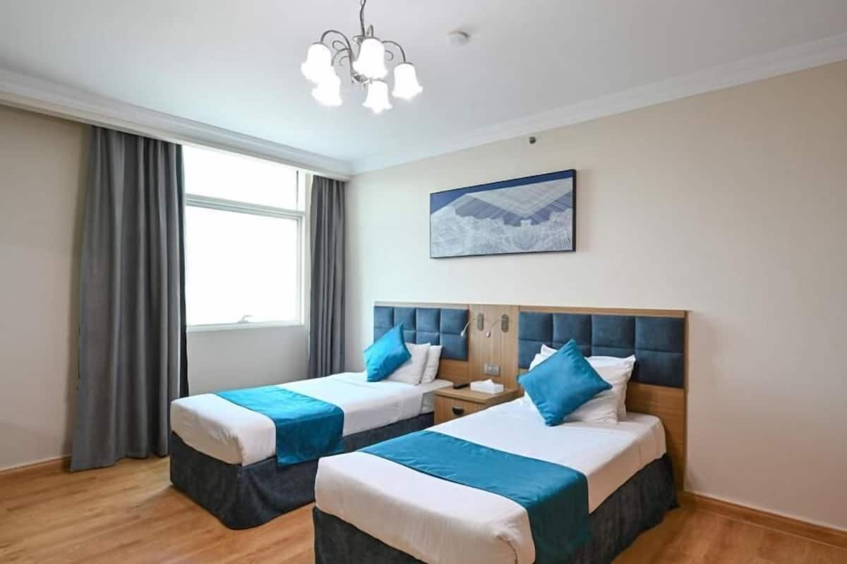 Habitación de hotel con dos camas, un candelabro, una ventana con cortinas y un cuadro en la pared.