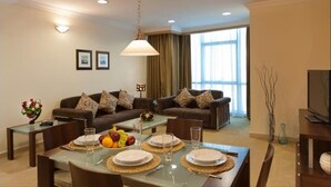 A furnished living room with a sofa set, a dining table set for four, and a television on a stand. - Saray Mushereib Hotel (Doha)