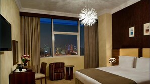 A hotel room with a large bed, a desk with a chair, a small table, a television, and a view of the cityscape at night. - Saray Mushereib Hotel (Doha)
