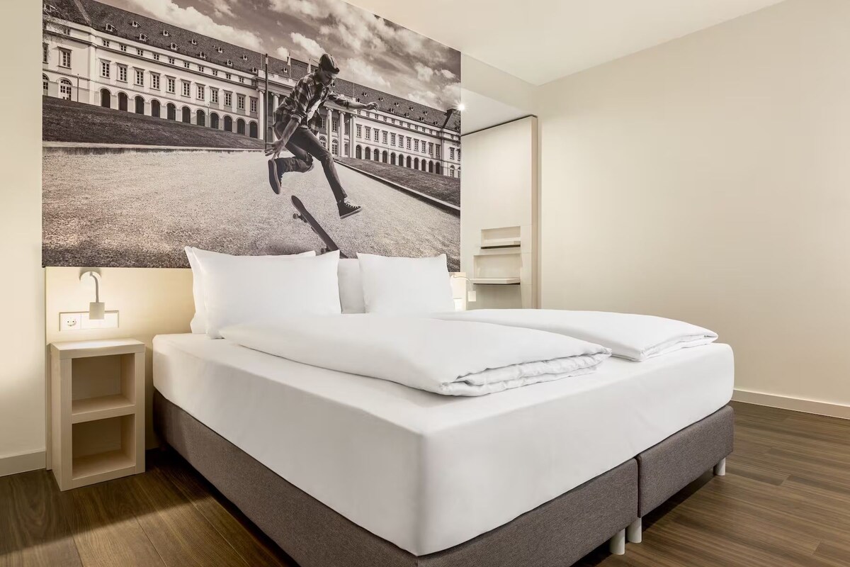 Habitación de hotel moderna con una cama grande, una mesita de noche, una lámpara fijada a la pared y un cuadro grande con una imagen de un edificio.
