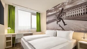Quarto de hotel com cama, escrivaninha, cadeira e uma grande janela com cortinas.