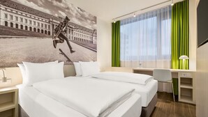 Quarto de hotel com cama, escrivaninha, cadeira, abajur e uma grande janela com cortinas verdes.