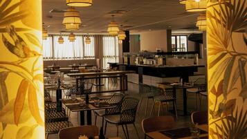 Un ristorante con una zona pranzo centrale, diverse luci a sospensione e una cucina visibile sullo sfondo.