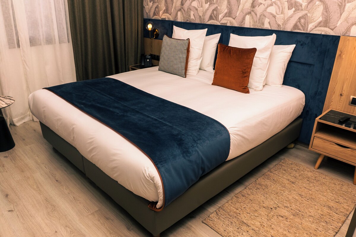 A large bed with a dark headboard, white bedding, and multiple pillows. There is a bedside table with a lamp, a small round table, and a wooden nightstand with a drawer.