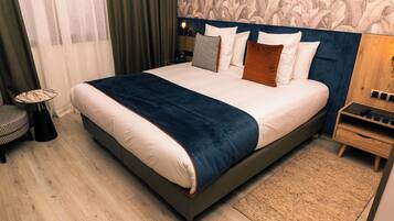 A large bed with a dark headboard, white bedding, and multiple pillows. There is a bedside table with a lamp, a small round table, and a wooden nightstand with a drawer.