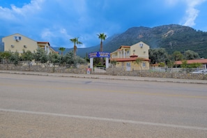 A hotel with a signboard, a paved road, and a mountainous backdrop. - Goldlion Hotel (Fethiye)