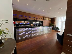 Zona de bar con mostrador de madera, un letrero que dice 'LYONS' y un área de estar con una planta y un sofá.