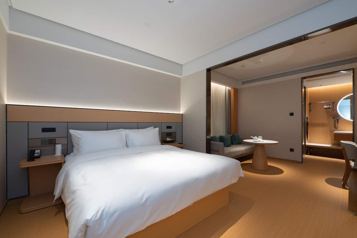 Quarto de hotel moderno com uma cama grande, mesas de cabeceira, um espaço de estar com uma mesa redonda e um banheiro visível por uma porta aberta.