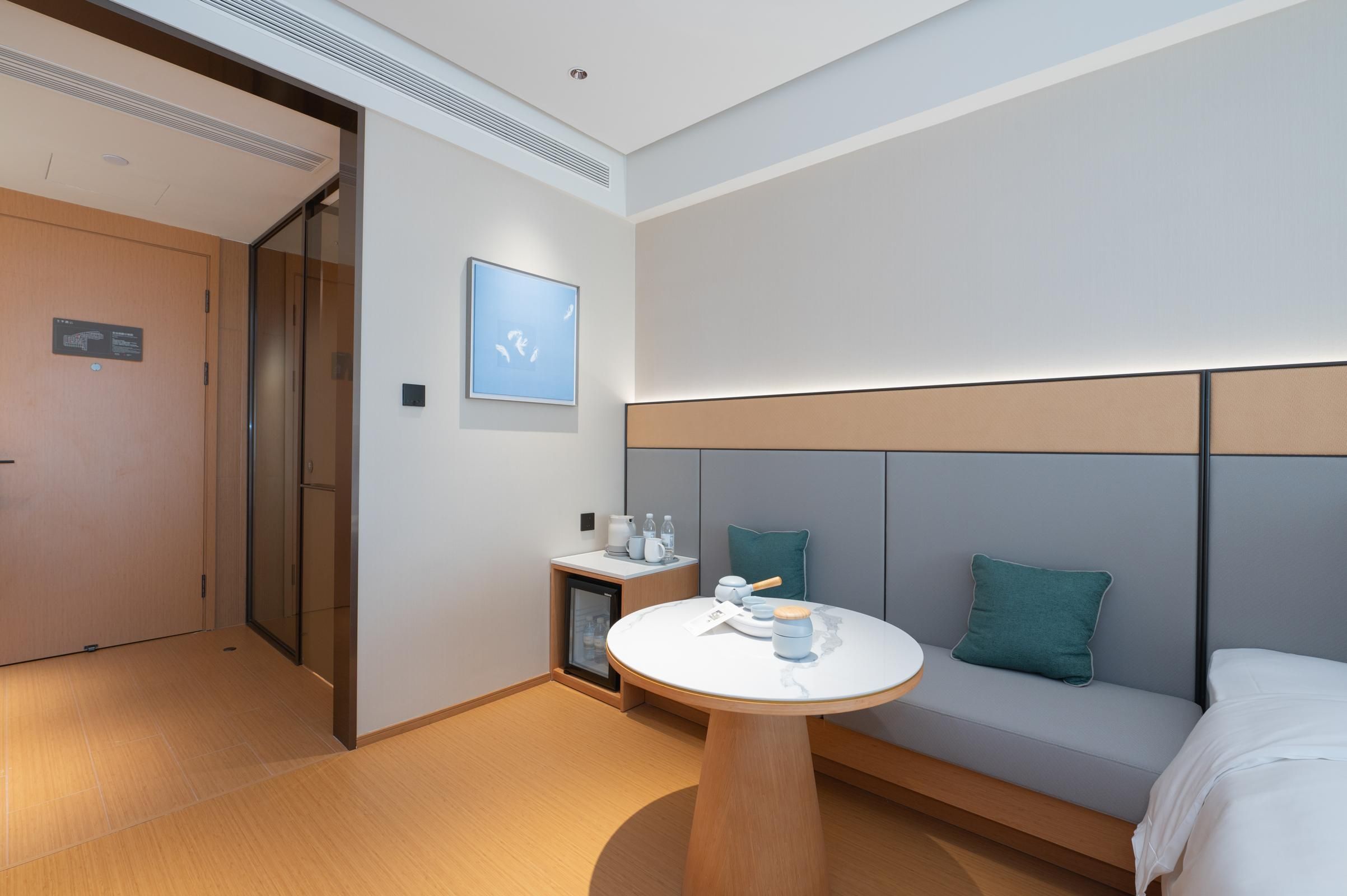 A modern hotel room with a sofa, a small table, a bedside table with a lamp, and a wall-mounted artwork.