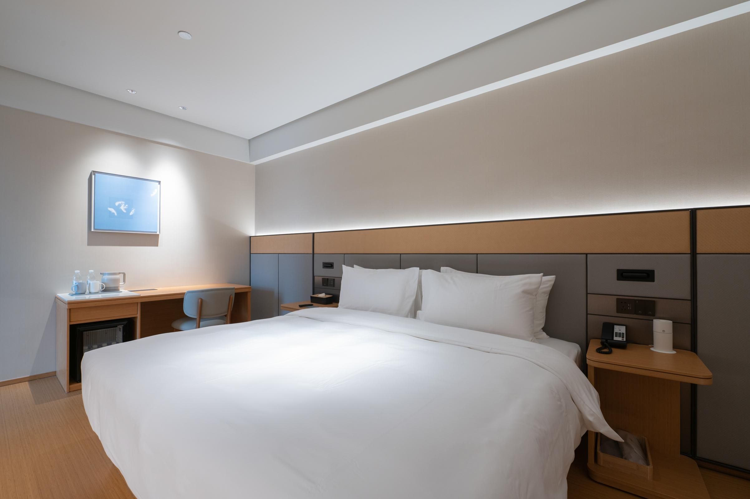 A modern hotel room with a large bed, a desk with a chair, a small table with a phone and a cup, and a wall-mounted artwork.