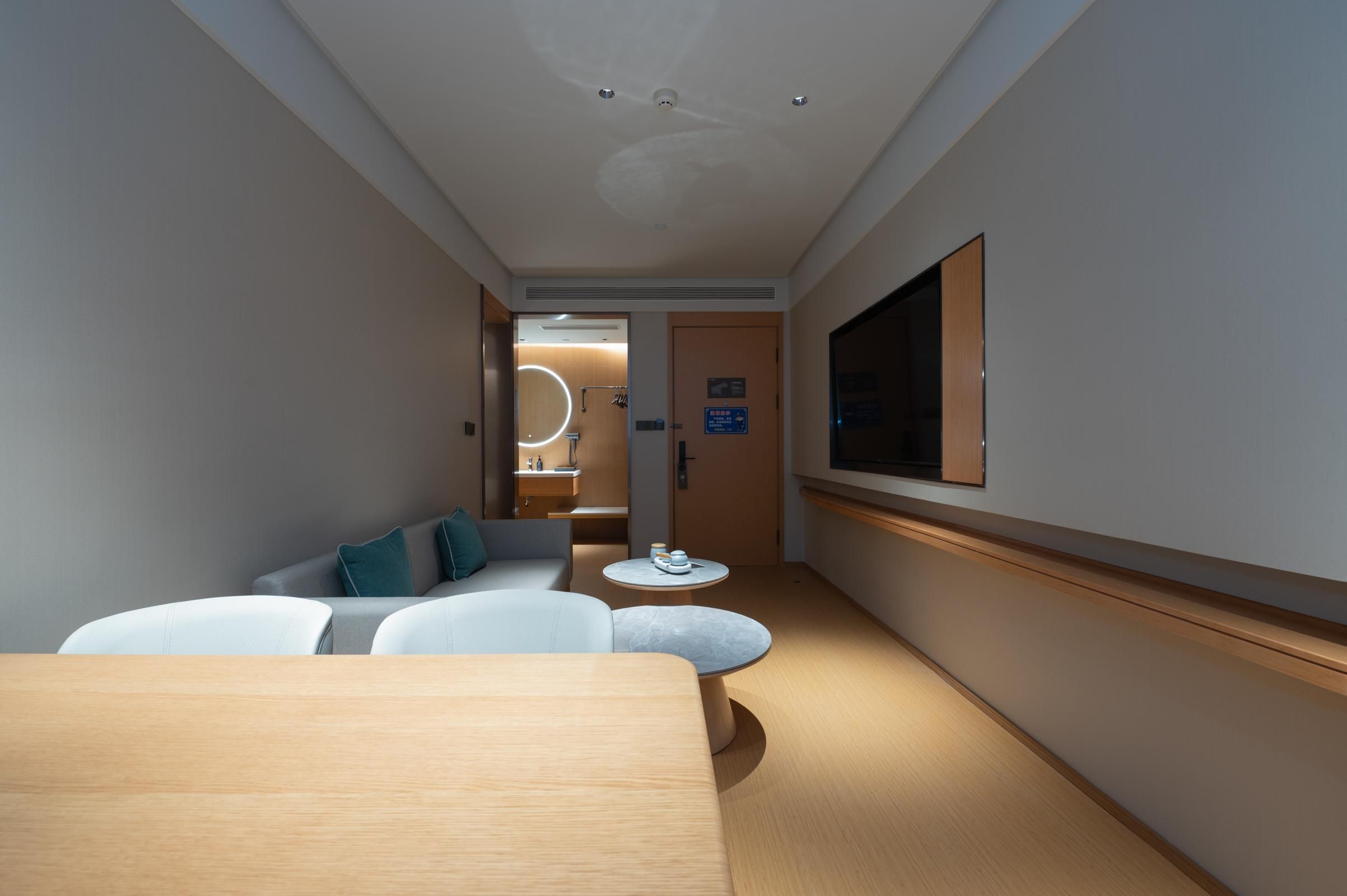 A hotel room with a wooden floor, a sofa, a small round table, a TV mounted on the wall, and a door leading to a bathroom.