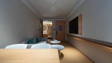 A hotel room with a wooden floor, a sofa, a small round table, a TV mounted on the wall, and a door leading to a bathroom.