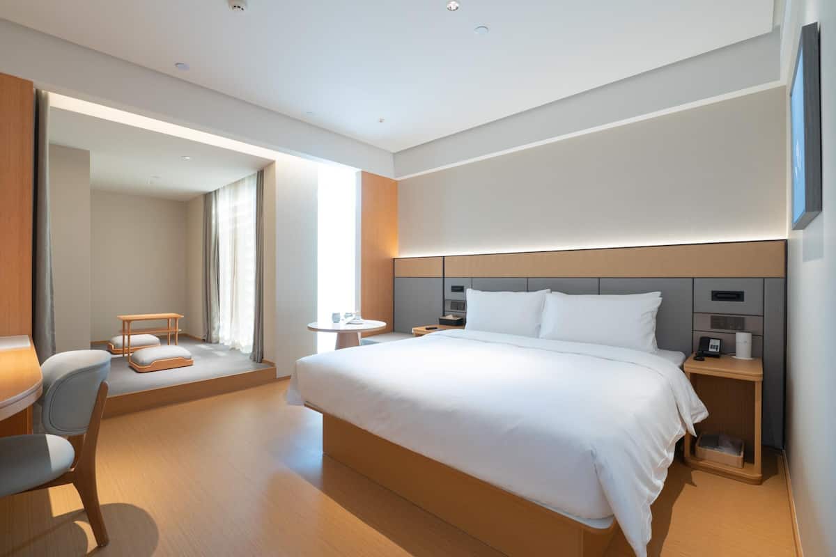 Quarto de hotel moderno com uma cama grande, uma escrivaninha, uma cadeira, uma mesinha de cabeceira e um criado-mudo.