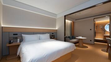 A modern hotel room with a large bed, bedside tables, a seating area with a round table, and a bathroom visible through an open door.