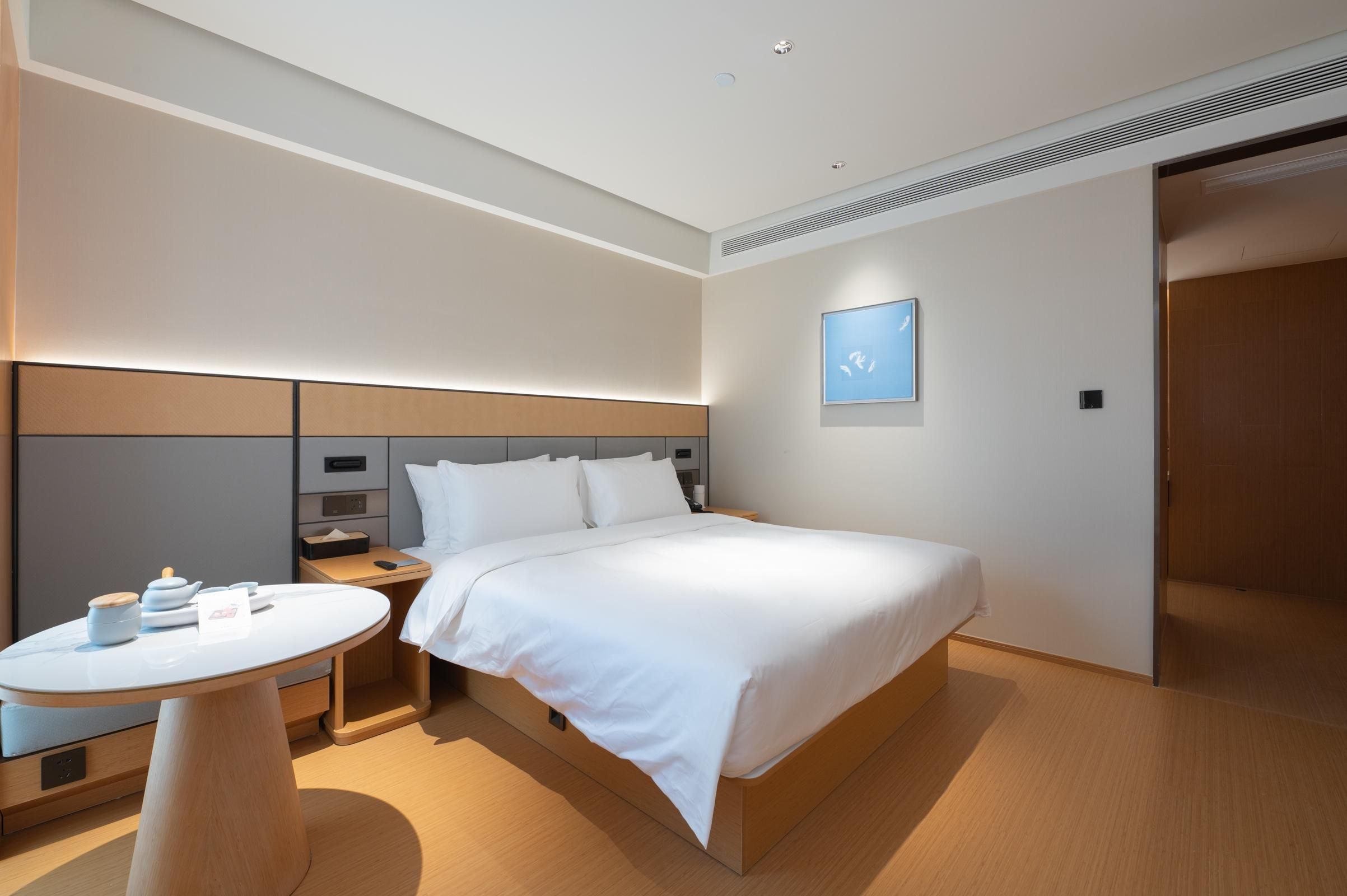 A modern hotel room with a bed, bedside tables, a small round table, and a wall-mounted light.