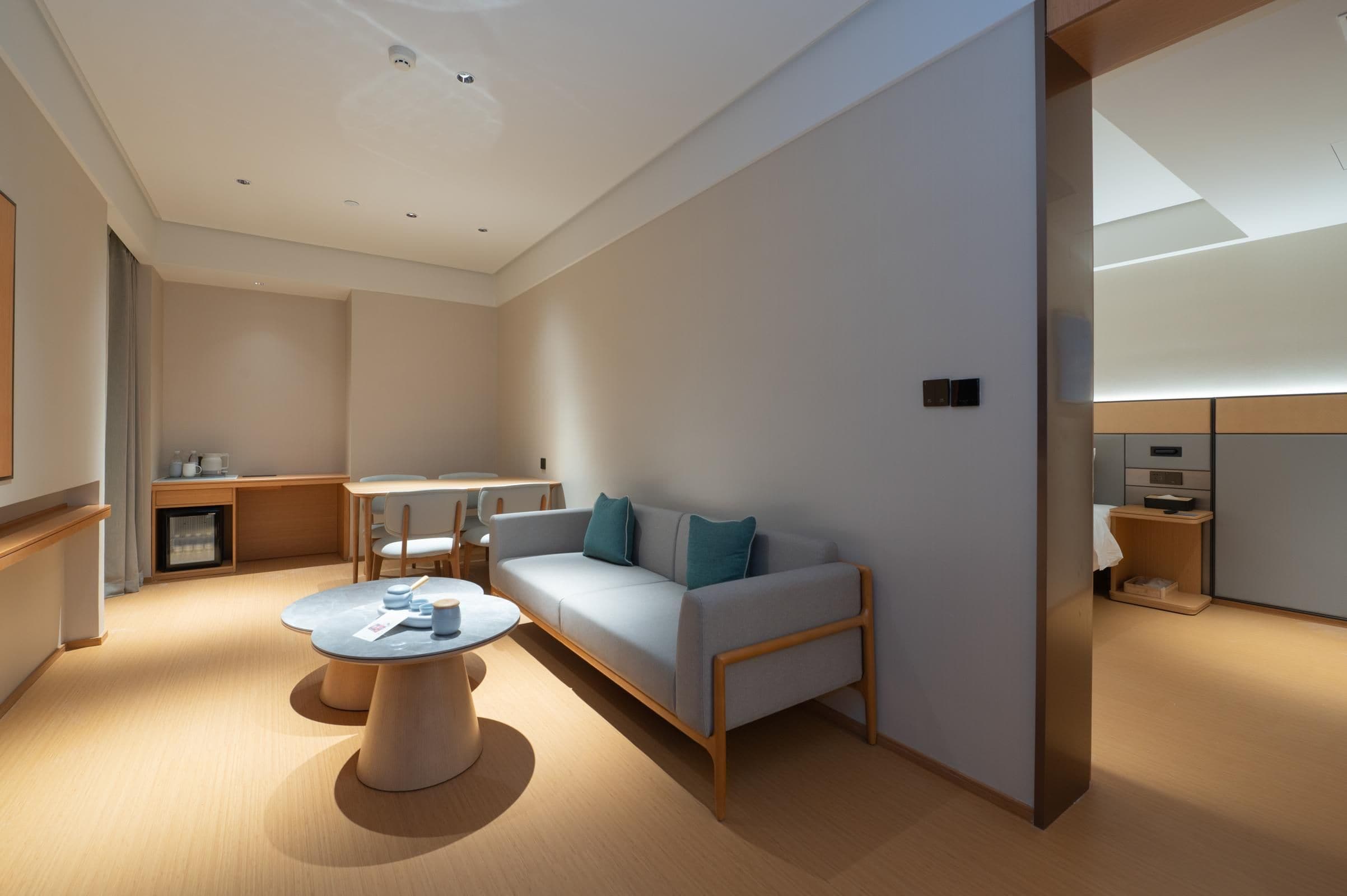 A modern hotel room with a sofa, a round coffee table, a desk, and a small kitchenette.
