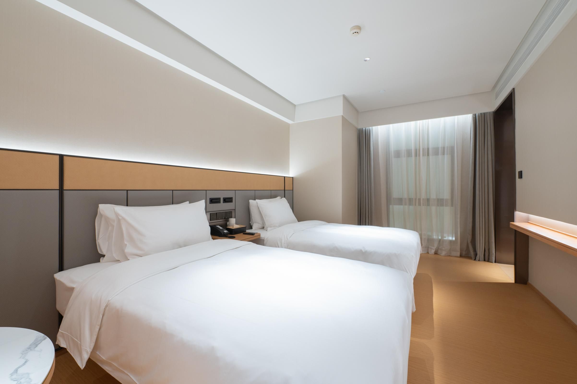 A hotel room with two beds, a bedside table, a desk, a chair, and a large window with curtains.