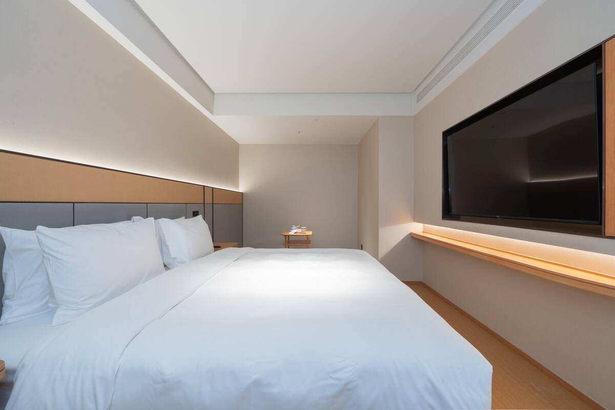 Quarto de hotel com uma cama grande, televisão fixada na parede e uma pequena mesa de cabeceira.