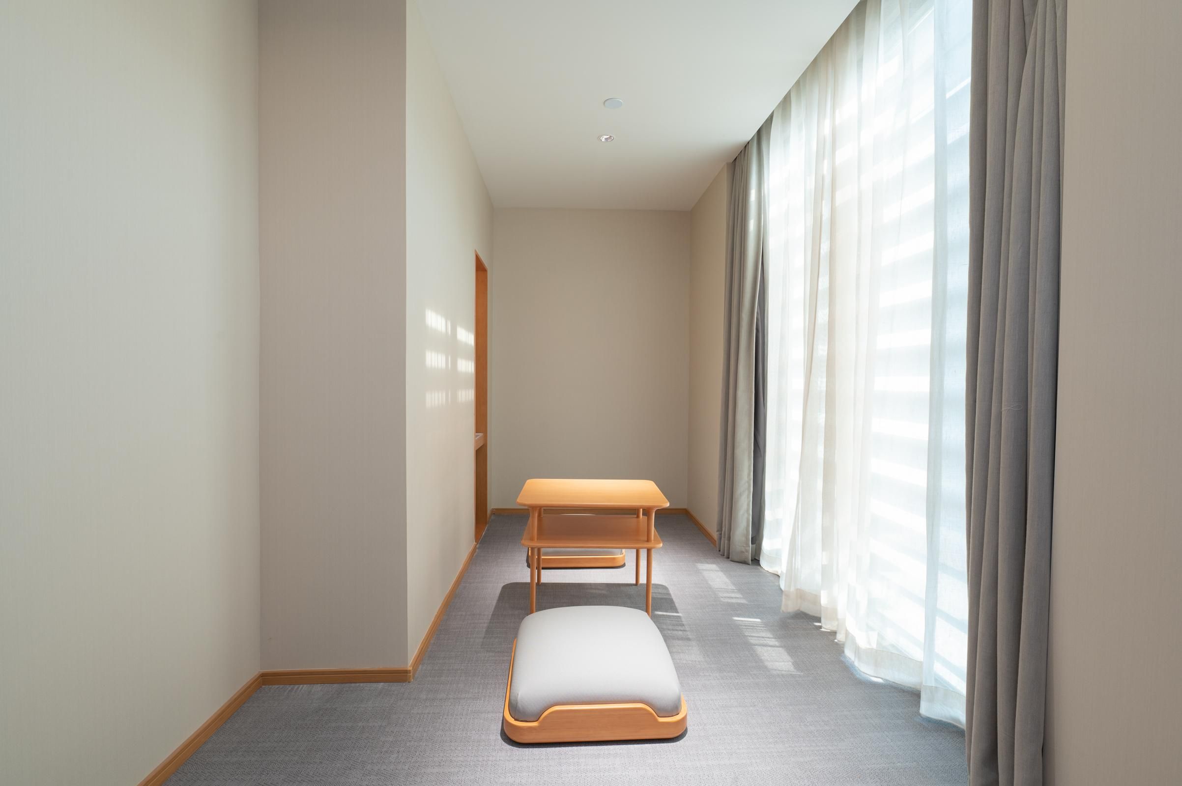 A narrow hotel corridor with a wooden bench and a small table, large windows with sheer curtains, and a carpeted floor.