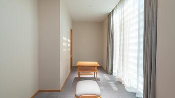 A narrow hotel corridor with a wooden bench and a small table, large windows with sheer curtains, and a carpeted floor.
