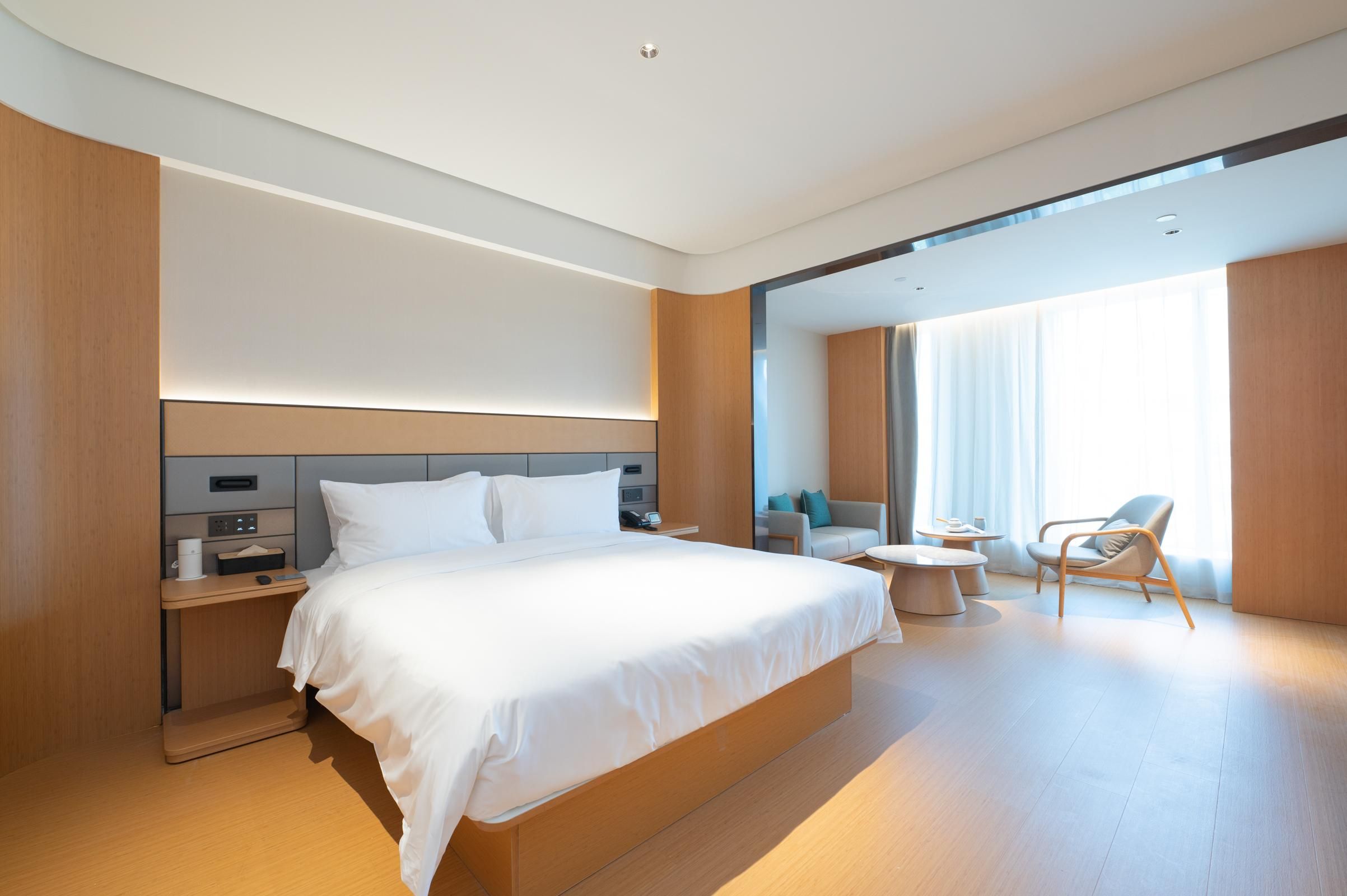 A modern hotel room with a large bed, bedside tables, a seating area with a chair and table, and a large window with curtains.