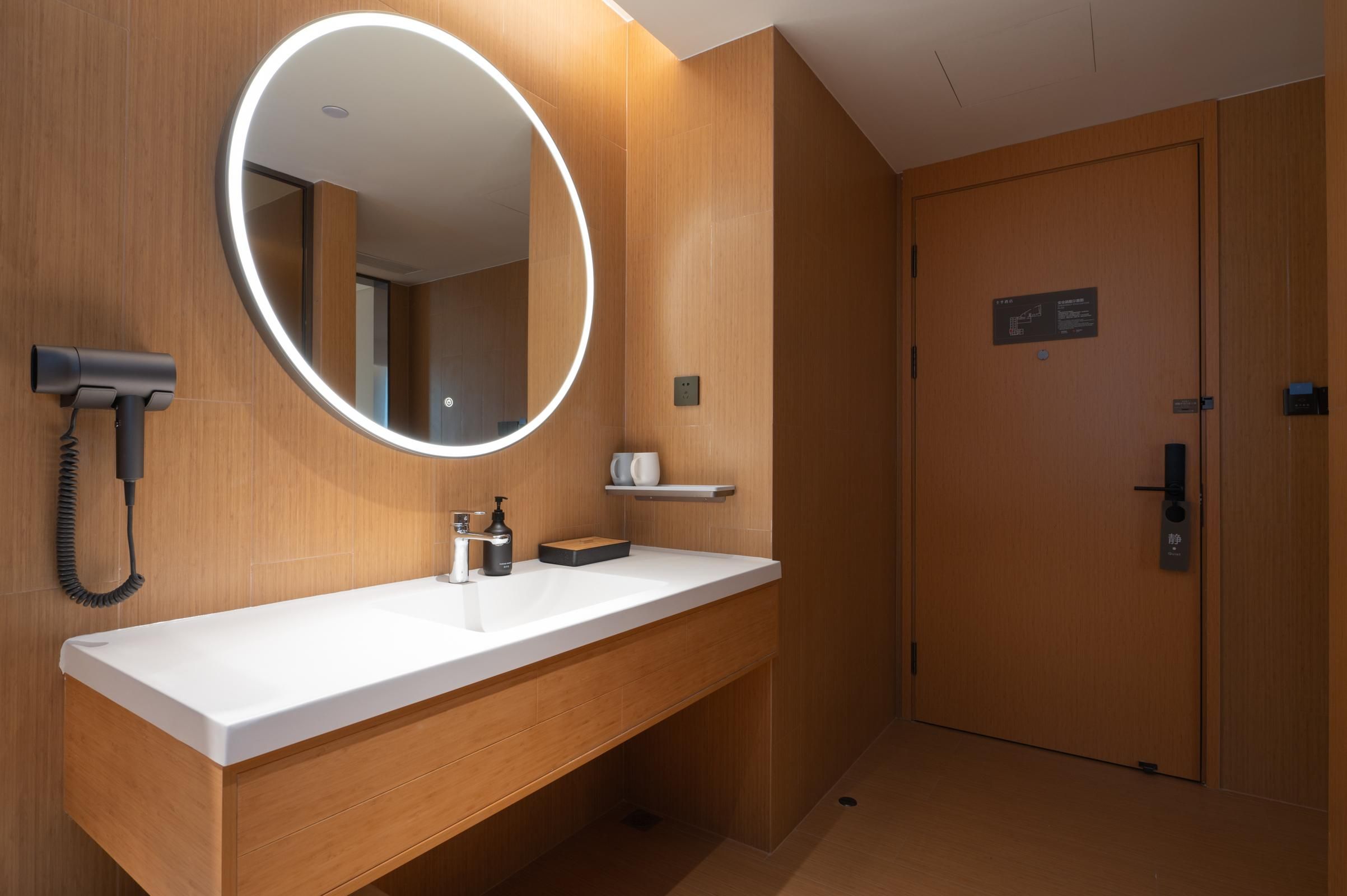 A modern bathroom with a white sink, a round mirror with lights, a hairdryer, and a wooden door.