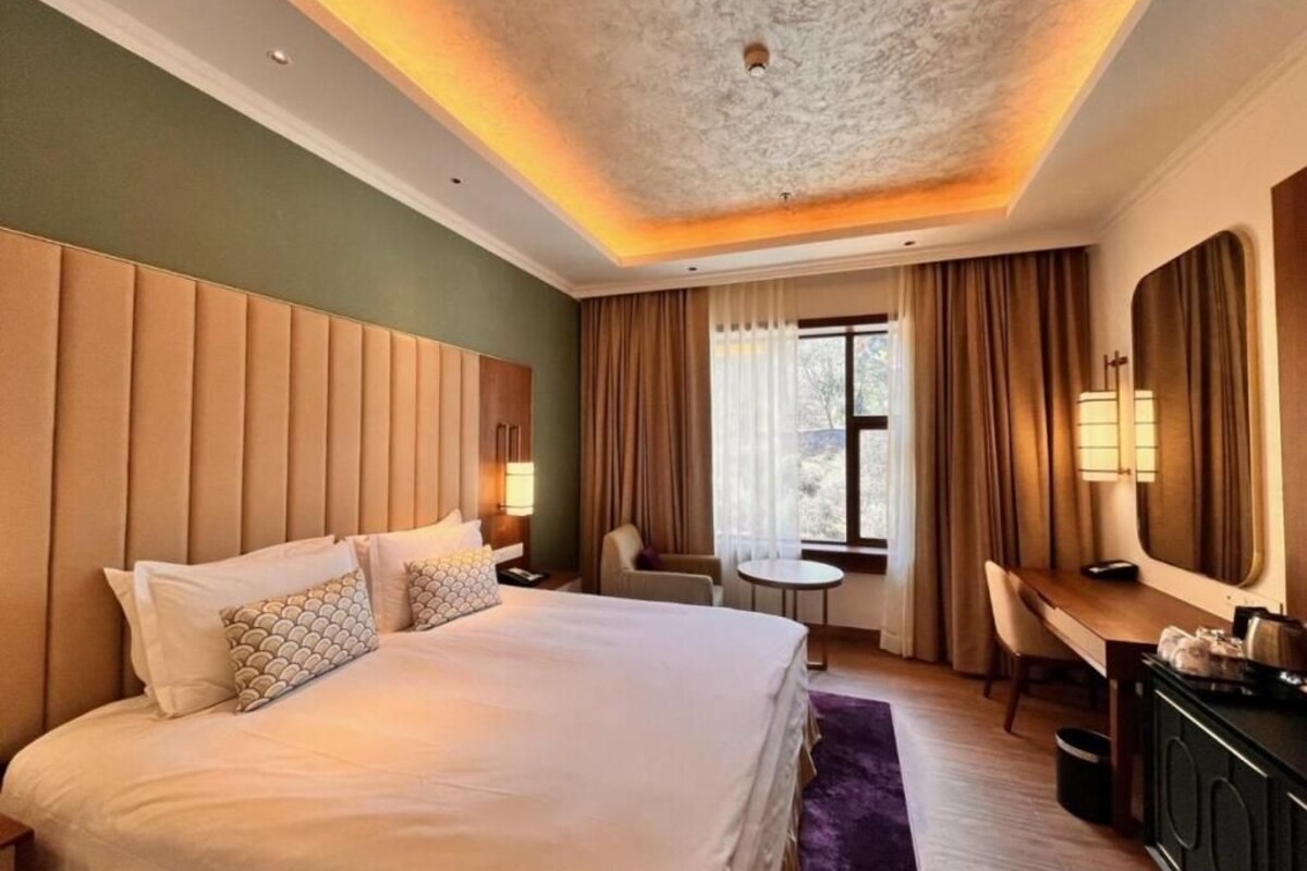 Quarto de hotel com uma cama grande, uma escrivaninha, uma cadeira, uma mesinha e uma janela com cortinas.