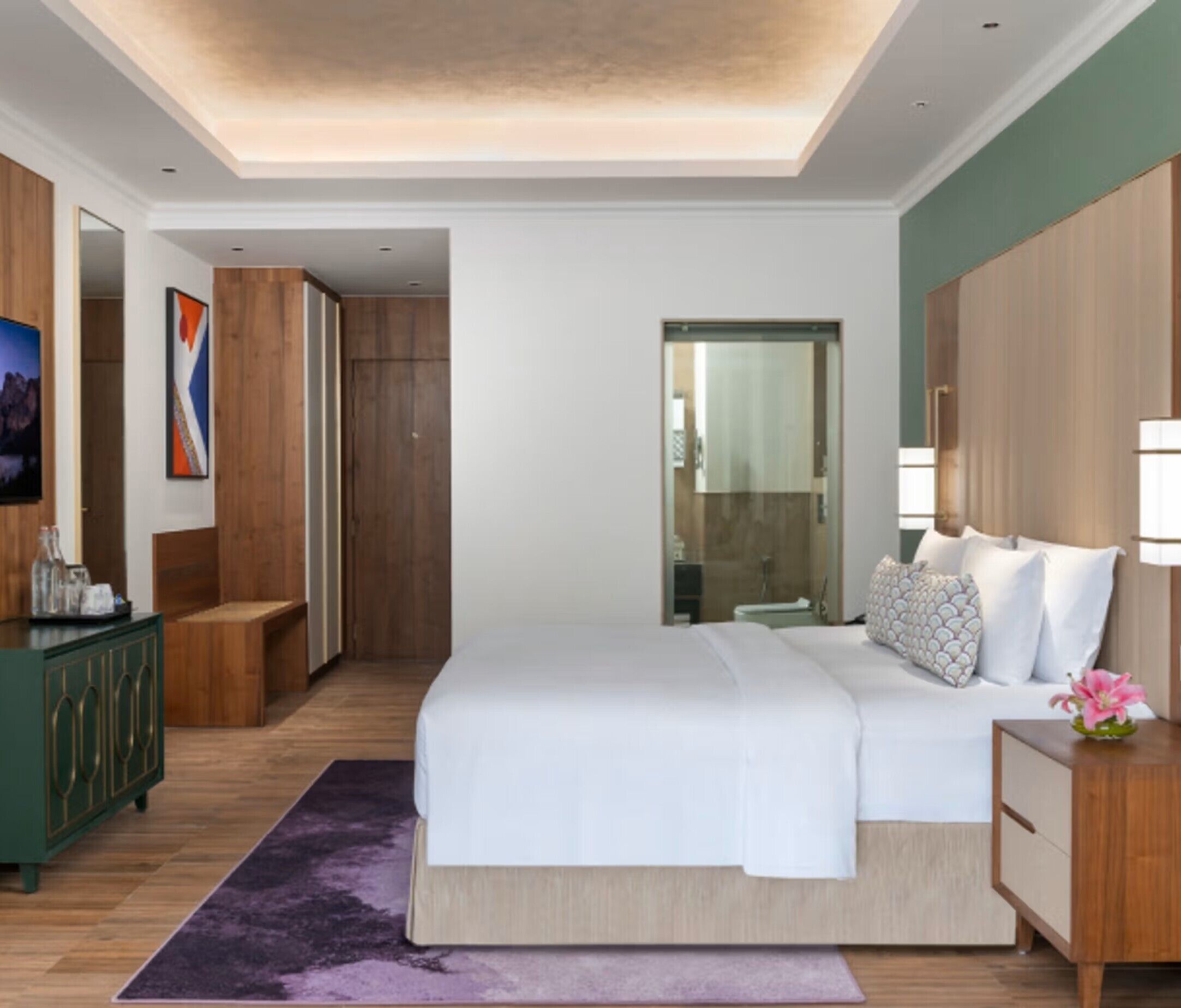 A modern hotel room with a large bed, wooden wardrobe, a green cabinet, and a bathroom visible through a glass door.