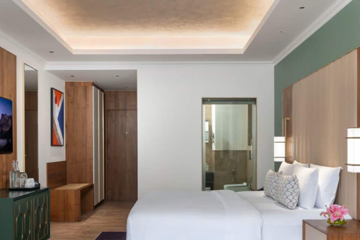 A modern hotel room with a large bed, wooden wardrobe, a green cabinet, and a bathroom visible through a glass door.