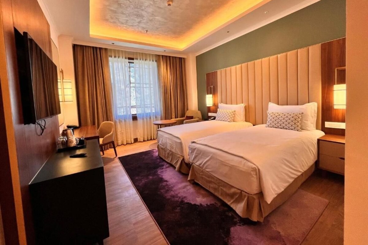 Quarto de hotel com duas camas, uma mesa, uma cadeira, uma televisão e uma janela grande com cortinas.