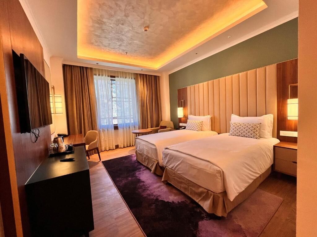A hotel room with two beds, a desk, a chair, a TV, and a large window with curtains.