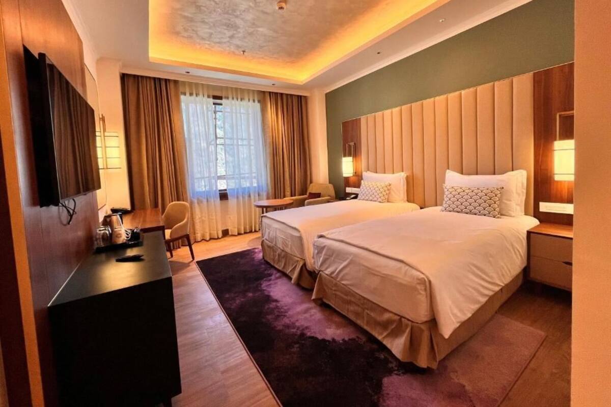 Ein Hotelzimmer mit zwei Betten, einem Schreibtisch, einem Sessel, einem Fernseher und einem großen Fenster mit Vorhängen.