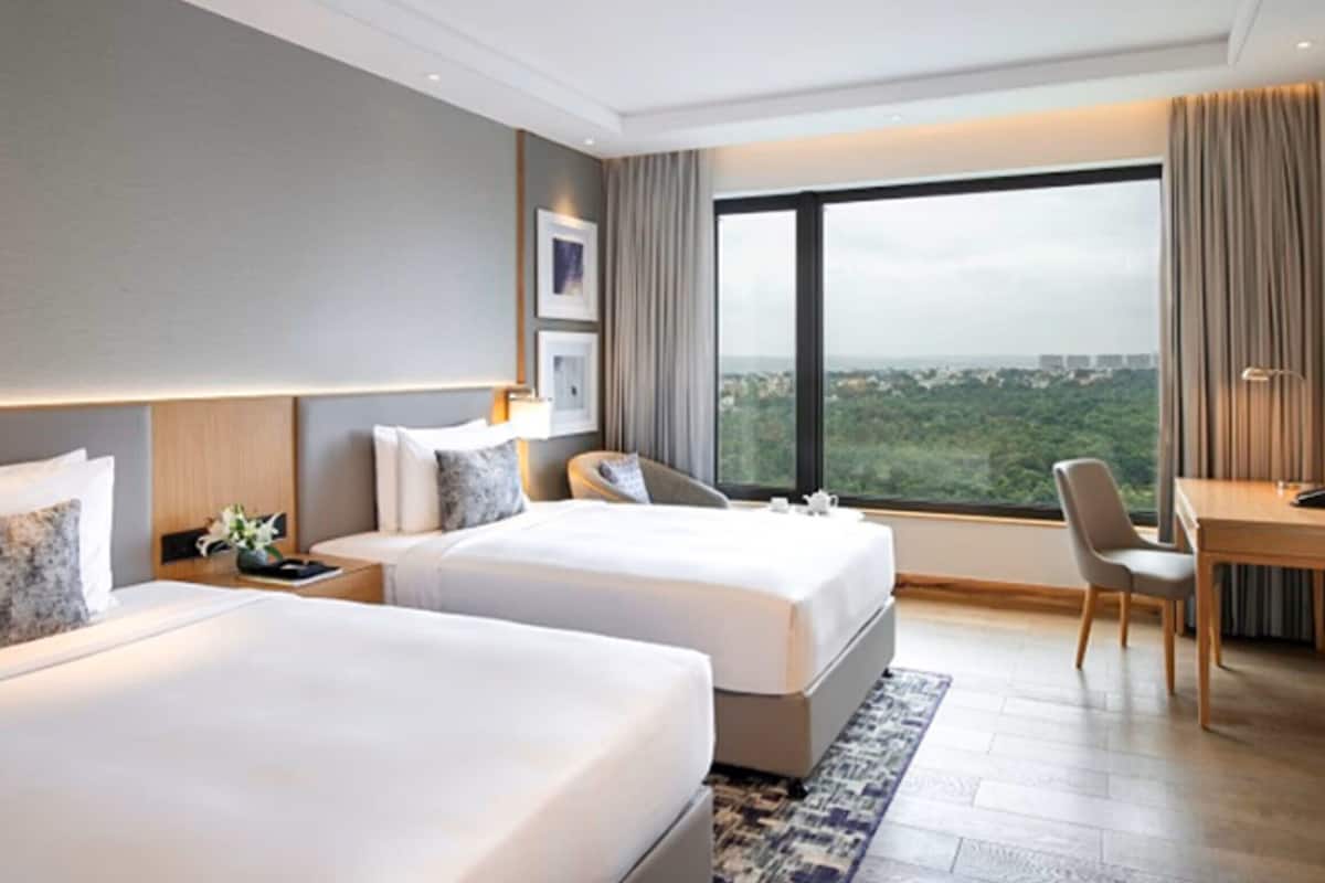 A hotel room with two beds, a desk, and a large window offering a city view.
