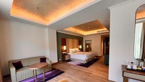 Quarto de hotel com cama, sofá, mesa redonda pequena, cadeira, espelho e piso de madeira.