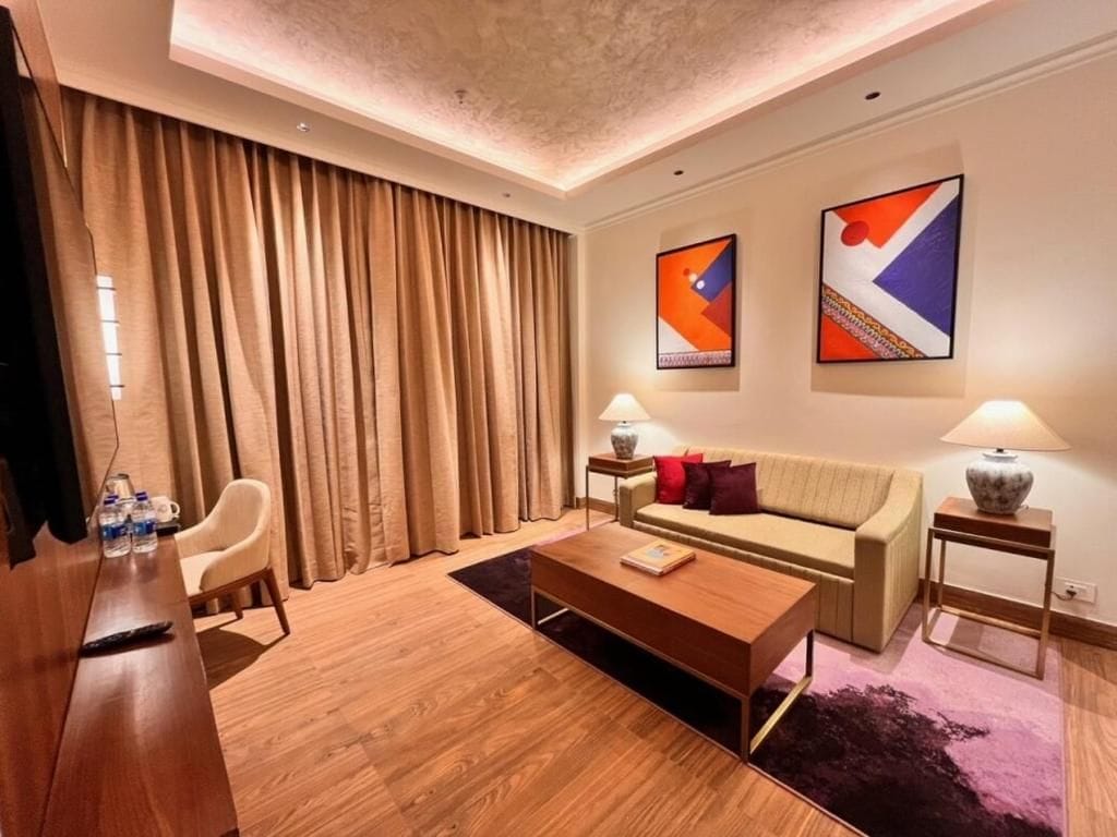 A modern hotel room with a wooden coffee table, a sofa, a TV, and a large window with curtains.