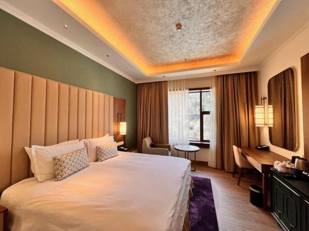 A hotel room with a large bed, a desk, a chair, a small table, and a window with curtains.